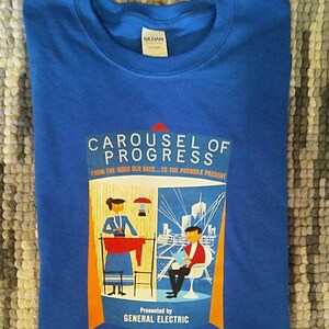K&ouml;nnte beinhalten: Ein blaues T-Shirt mit einer Grafik im Vintage-Stil eines Karussells mit dem Text "Carousel of Progress" und "From the good old days... to the possible present". Die Grafik zeigt auch eine Frau in einem blauen Kleid und einen Mann in einem roten Hemd. Der Text "Presented by General Electric" befindet sich am unteren Rand der Grafik.