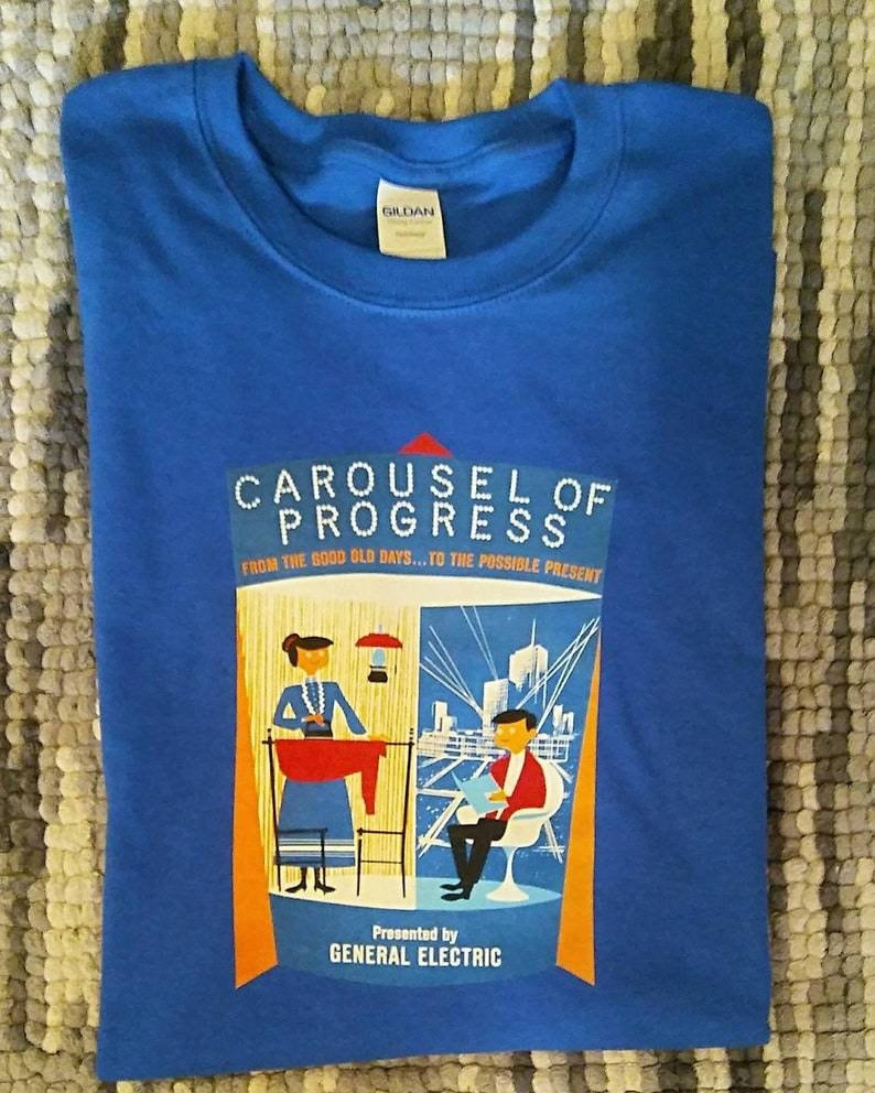 K&ouml;nnte beinhalten: Ein blaues T-Shirt mit einer Grafik im Vintage-Stil eines Karussells mit dem Text "Carousel of Progress" und "From the good old days... to the possible present". Die Grafik zeigt auch eine Frau in einem blauen Kleid und einen Mann in einem roten Hemd. Der Text "Presented by General Electric" befindet sich am unteren Rand der Grafik.