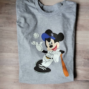 May include: A grey t-shirt featuring a cartoon image of Mickey Mouse wearing a baseball cap and uniform. The cap has the letters "GR" on it. Mickey is holding a baseball bat and a baseball is in the background.