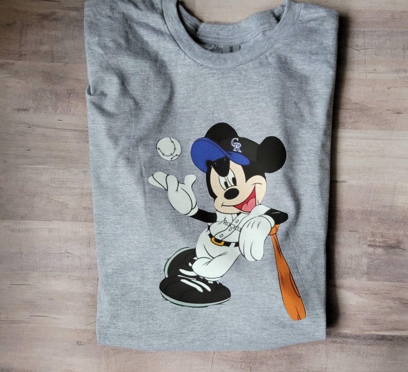 May include: A grey t-shirt featuring a cartoon image of Mickey Mouse wearing a baseball cap and uniform. The cap has the letters "GR" on it. Mickey is holding a baseball bat and a baseball is in the background.
