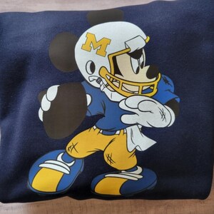 May include: A navy blue sweatshirt with a cartoon image of Mickey Mouse wearing a football helmet with the letter "M" on it. Mickey is holding a football and wearing a blue and yellow football uniform.