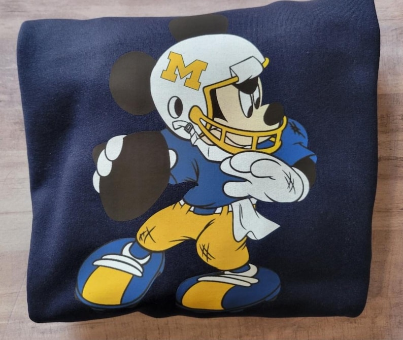 May include: A navy blue sweatshirt with a cartoon image of Mickey Mouse wearing a football helmet with the letter "M" on it. Mickey is holding a football and wearing a blue and yellow football uniform.