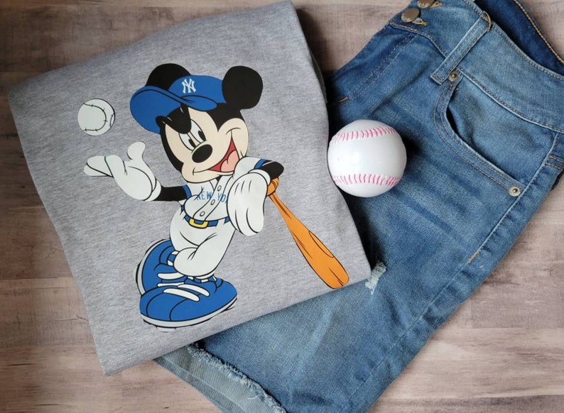 May include: A grey sweatshirt featuring a cartoon image of Mickey Mouse wearing a baseball uniform and holding a bat. The image includes the text "NEW YORK" on the uniform. A baseball and a pair of blue jeans are also in the image.