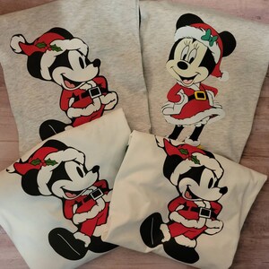 K&ouml;nnte beinhalten: Vier Sweatshirts mit Mickey und Minnie Mouse in Weihnachtskleidung. Mickey tr&auml;gt einen Weihnachtsmann-Anzug und -Hut mit Stechpalme. Minnie tr&auml;gt ein rotes Kleid und einen Weihnachtsmann-Hut mit gr&uuml;ner Schleife. Zwei Sweatshirts sind grau, zwei sind wei&szlig;.