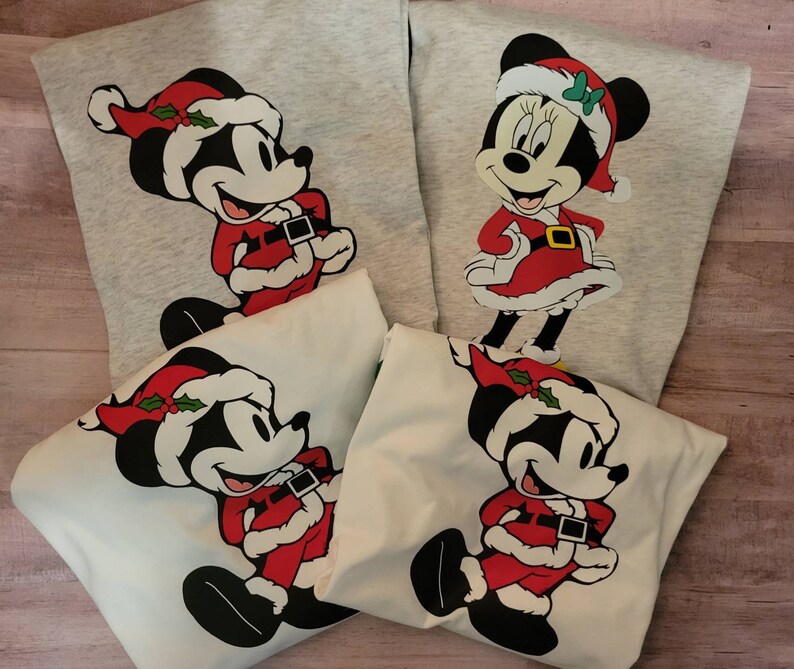 K&ouml;nnte beinhalten: Vier Sweatshirts mit Mickey und Minnie Mouse in Weihnachtskleidung. Mickey tr&auml;gt einen Weihnachtsmann-Anzug und -Hut mit Stechpalme. Minnie tr&auml;gt ein rotes Kleid und einen Weihnachtsmann-Hut mit gr&uuml;ner Schleife. Zwei Sweatshirts sind grau, zwei sind wei&szlig;.