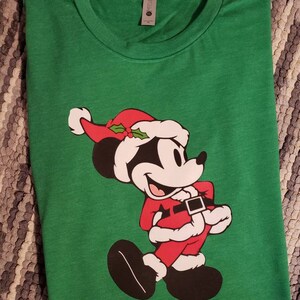 K&ouml;nnte beinhalten: Ein gr&uuml;nes T-Shirt mit einer Cartoonfigur in einem Weihnachtsmann-Kost&uuml;m. Die Figur tr&auml;gt einen roten Weihnachtsmann-Anzug, Hut und schwarze Schuhe. Das T-Shirt ist gefaltet und hat ein Etikett am Kragen.