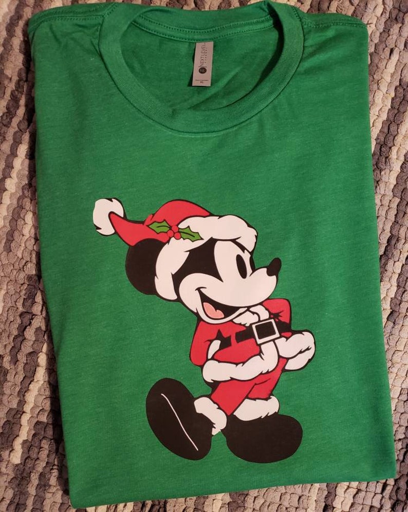 K&ouml;nnte beinhalten: Ein gr&uuml;nes T-Shirt mit einer Cartoonfigur in einem Weihnachtsmann-Kost&uuml;m. Die Figur tr&auml;gt einen roten Weihnachtsmann-Anzug, Hut und schwarze Schuhe. Das T-Shirt ist gefaltet und hat ein Etikett am Kragen.