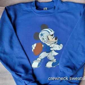 May include: Blue crewneck sweatshirt featuring a graphic of Mickey Mouse wearing a Dallas Cowboys football uniform and holding a football. The text "crewneck sweatshirt" is printed at the bottom of the sweatshirt.