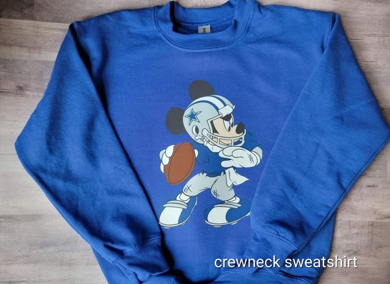 May include: Blue crewneck sweatshirt featuring a graphic of Mickey Mouse wearing a Dallas Cowboys football uniform and holding a football. The text "crewneck sweatshirt" is printed at the bottom of the sweatshirt.