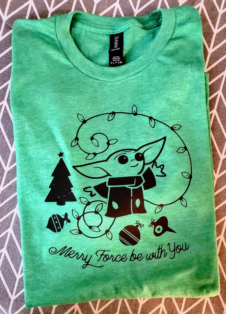 May include: Green t-shirt with a black and white graphic of a character from Star Wars, a Christmas tree, and ornaments. The text "Merry Force be with You" is printed below the graphic.
