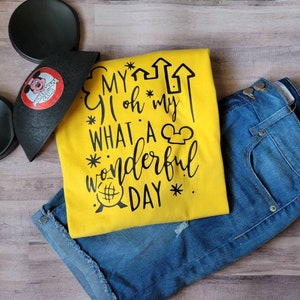 May include: A yellow t-shirt with black text that reads "My oh my what a wonderful day" with a Mickey Mouse head graphic. The shirt is paired with blue denim shorts and a black Mickey Mouse ear headband.