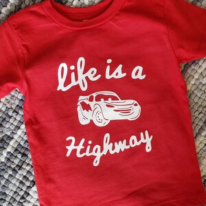 K&ouml;nnte beinhalten: Rotes T-Shirt mit kurzen &Auml;rmeln und einem wei&szlig;en Aufdruck eines roten Rennwagens und dem Text "life is a highway".