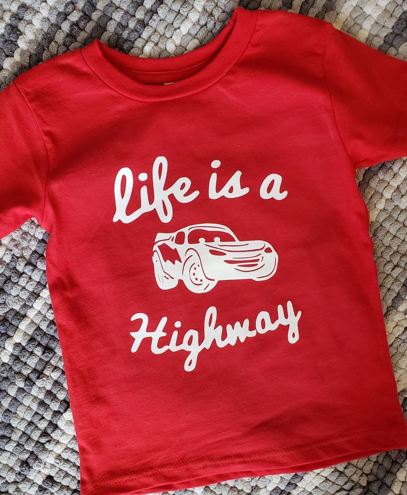 K&ouml;nnte beinhalten: Rotes T-Shirt mit kurzen &Auml;rmeln und einem wei&szlig;en Aufdruck eines roten Rennwagens und dem Text "life is a highway".