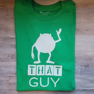 May include: Green t-shirt with a white silhouette of a monster from the movie Monsters Inc. The text "THAT GUY" is printed below the monster.