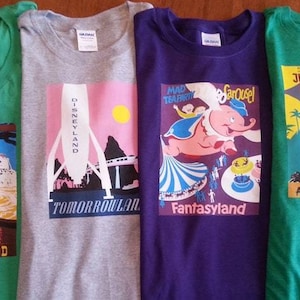 K&ouml;nnte beinhalten: Vier T-Shirts mit verschiedenen Disney-Park-Designs. Die Shirts sind gr&uuml;n, grau, lila und gr&uuml;n. Die Designs sind Frontierland, Tomorrowland, Fantasyland und Adventureland.