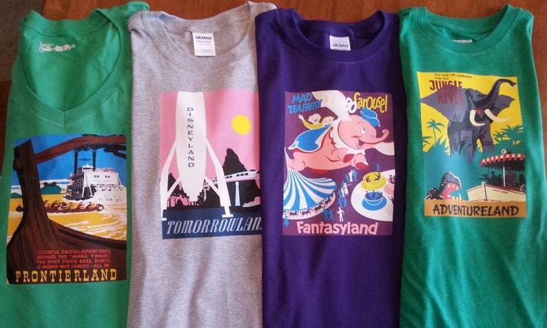 K&ouml;nnte beinhalten: Vier T-Shirts mit verschiedenen Disney-Park-Designs. Die Shirts sind gr&uuml;n, grau, lila und gr&uuml;n. Die Designs sind Frontierland, Tomorrowland, Fantasyland und Adventureland.