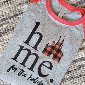 Op de afbeelding: Een grijs en rood raglan t-shirt met zwarte tekst die "home for the holidays" luidt en een zwarte en rode geruite kasteelgrafiek.