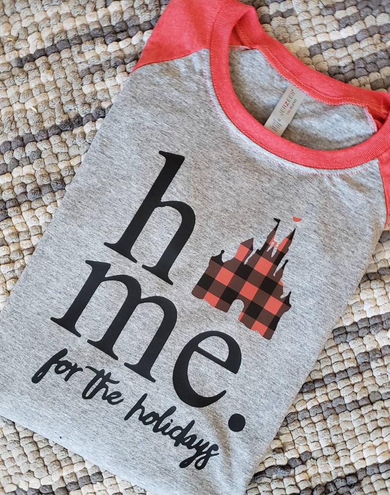 Op de afbeelding: Een grijs en rood raglan t-shirt met zwarte tekst die "home for the holidays" luidt en een zwarte en rode geruite kasteelgrafiek.