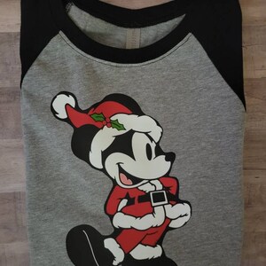 K&ouml;nnte beinhalten: Graues Raglan-Shirt mit schwarzen &Auml;rmeln und einem schwarzen Kragen. Das Shirt zeigt eine Cartoonfigur in einem Weihnachtsmann-Outfit, komplett mit rotem Hut, Mantel und schwarzen Schuhen. Die Figur l&auml;chelt.