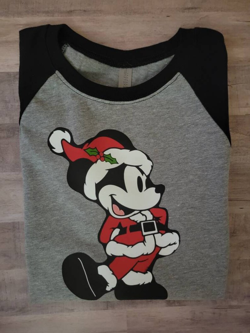 K&ouml;nnte beinhalten: Graues Raglan-Shirt mit schwarzen &Auml;rmeln und einem schwarzen Kragen. Das Shirt zeigt eine Cartoonfigur in einem Weihnachtsmann-Outfit, komplett mit rotem Hut, Mantel und schwarzen Schuhen. Die Figur l&auml;chelt.