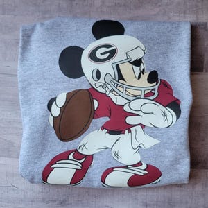 May include: A gray t-shirt with a cartoon image of Mickey Mouse dressed as a football player. Mickey is wearing a red jersey, white pants, a white helmet with a red and black 'G' on it, and a brown football.