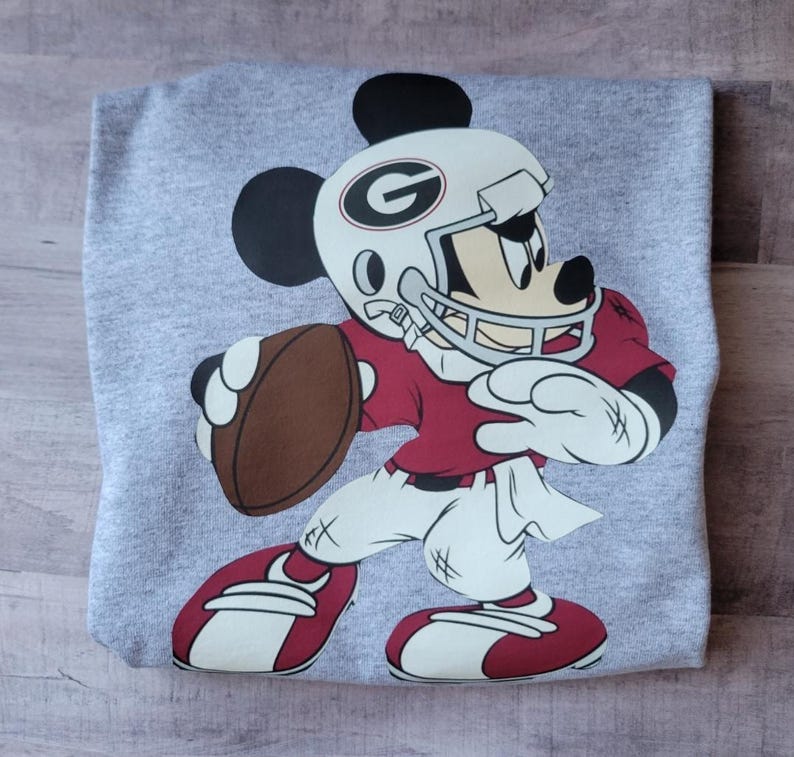 May include: A gray t-shirt with a cartoon image of Mickey Mouse dressed as a football player. Mickey is wearing a red jersey, white pants, a white helmet with a red and black 'G' on it, and a brown football.