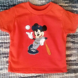 May include: Red short-sleeved t-shirt featuring a cartoon image of Mickey Mouse playing baseball. Mickey is wearing a baseball uniform with a Detroit Tigers logo on his hat.
