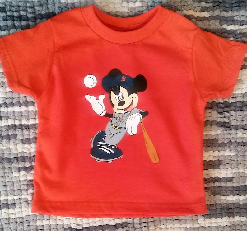 May include: Red short-sleeved t-shirt featuring a cartoon image of Mickey Mouse playing baseball. Mickey is wearing a baseball uniform with a Detroit Tigers logo on his hat.