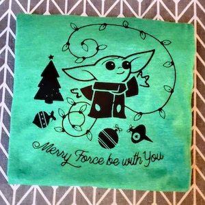 May include: A green t-shirt with a black design of a baby Yoda character wearing a Santa hat and holding a Christmas ornament. The design also includes a Christmas tree, ornaments, and a string of lights. The text "Merry Force be with You" is printed below the design.