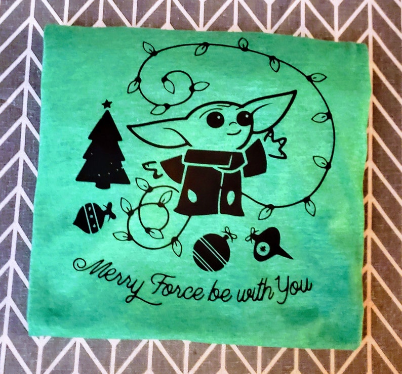 May include: A green t-shirt with a black design of a baby Yoda character wearing a Santa hat and holding a Christmas ornament. The design also includes a Christmas tree, ornaments, and a string of lights. The text "Merry Force be with You" is printed below the design.
