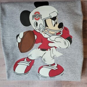 May include: A gray t-shirt with a cartoon image of Mickey Mouse dressed as a football player. Mickey is wearing a red and white jersey, a white helmet with the words "Ohio State" on it, and a brown football.
