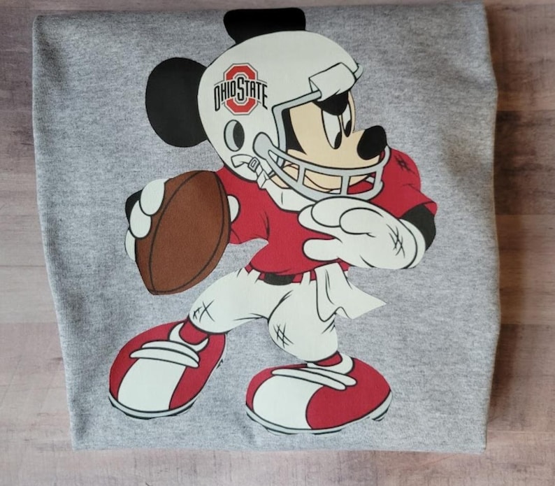 May include: A gray t-shirt with a cartoon image of Mickey Mouse dressed as a football player. Mickey is wearing a red and white jersey, a white helmet with the words "Ohio State" on it, and a brown football.