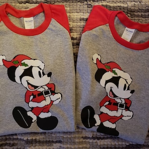 K&ouml;nnte beinhalten: Zwei graue Raglan-Shirts mit roten &Auml;rmeln und Kragen. Jedes Shirt zeigt eine Cartoon-Mickey-Mouse in einem Weihnachtsmann-Outfit, komplett mit rotem Hut, Anzug und schwarzen Schuhen. Die Shirts sind f&uuml;r die Weihnachtszeit.