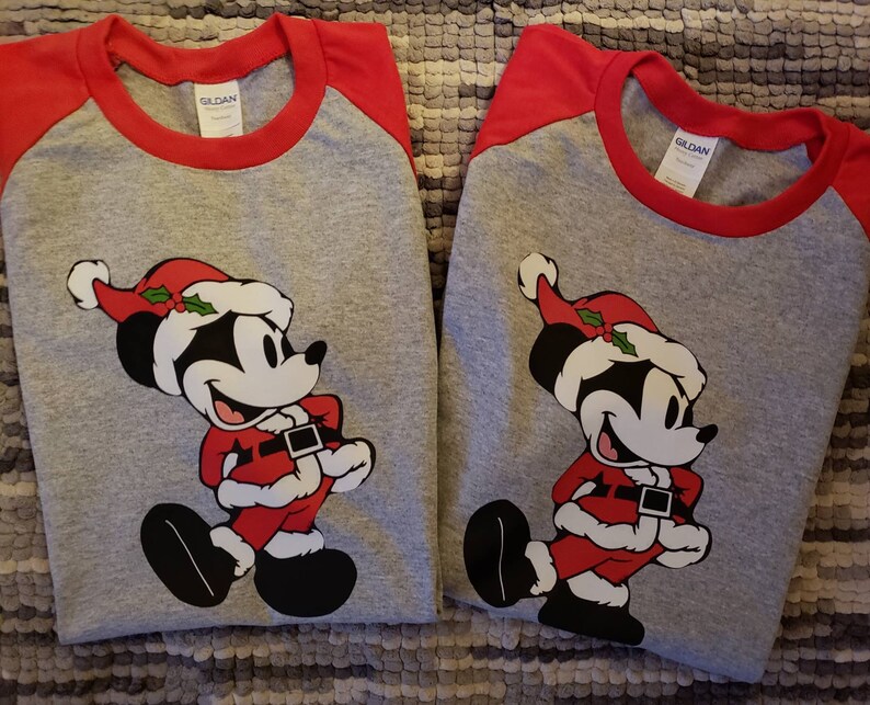 K&ouml;nnte beinhalten: Zwei graue Raglan-Shirts mit roten &Auml;rmeln und Kragen. Jedes Shirt zeigt eine Cartoon-Mickey-Mouse in einem Weihnachtsmann-Outfit, komplett mit rotem Hut, Anzug und schwarzen Schuhen. Die Shirts sind f&uuml;r die Weihnachtszeit.