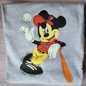 May include: A grey sweatshirt featuring a cartoon image of Mickey Mouse wearing a red baseball cap, a red and white baseball jersey with the word "Orioles" on it, yellow trousers, and black baseball boots. Mickey is holding a brown baseball bat and a baseball is in the air above him.