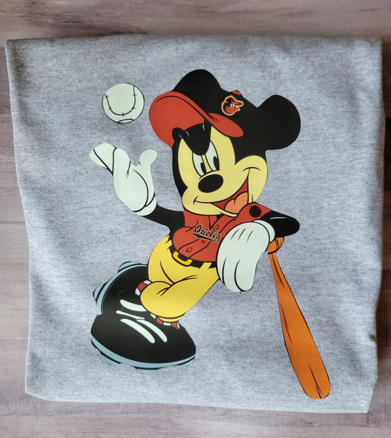 May include: A grey sweatshirt featuring a cartoon image of Mickey Mouse wearing a red baseball cap, a red and white baseball jersey with the word "Orioles" on it, yellow trousers, and black baseball boots. Mickey is holding a brown baseball bat and a baseball is in the air above him.