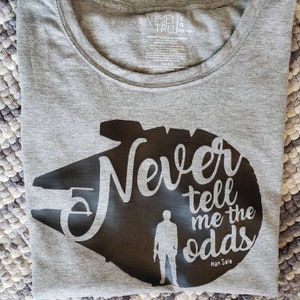 May include: A gray t-shirt with a black silhouette of the Millennium Falcon spaceship. The text "Never tell me the odds - Han Solo" is printed on the shirt.