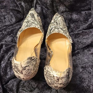Puede incluir: Un par de zapatos de tacón con punta afilada y estampado de piel de serpiente en tonos negro, blanco y gris. Los zapatos cuentan con paneles laterales transparentes y un forro interior color canela, con un diseño elegante y talón cerrado.
