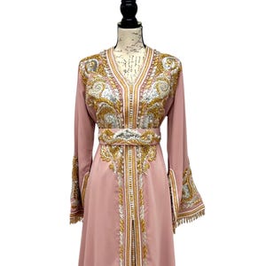 May include: A long, blush pink dress with intricate gold and silver embroidery. The dress features a V-neck, long sleeves with flared cuffs, and a matching embellished belt. The dress is displayed on a mannequin.