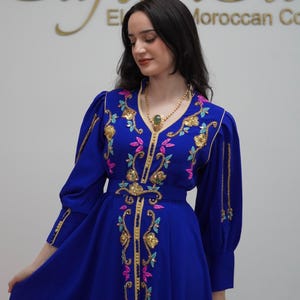 May include: A royal blue caftan dress with gold embroidery and colorful floral accents. The dress features long sleeves and a V-neckline, and is worn with a gold necklace with a green pendant. The caftan is from the "Caftan Sto" Moroccan collection.