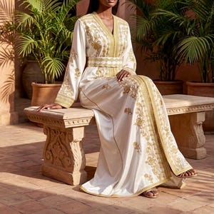 May include: A white caftan with gold embroidery and trim. The long-sleeved garment features a V-neck, a matching belt, and ornate floral patterns. The person is seated on a stone bench in an outdoor setting.