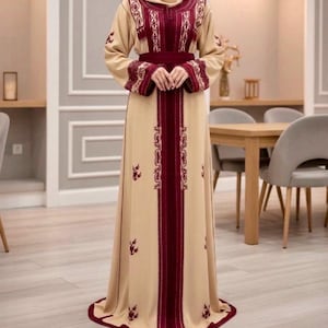 May include: A beige maxi dress with burgundy embroidery and accents. The dress features long sleeves with embroidered cuffs, a burgundy belt, and a central burgundy panel with ornate detailing. The dress is floor-length and suitable for special occasions.
