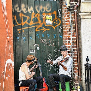 Può includere: Due musicisti di strada si esibiscono in una strada della città. Un musicista suona il violino mentre l'altro suona la chitarra. I musicisti indossano cappelli e sono in piedi davanti a un muro di mattoni con graffiti.