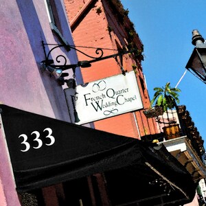 May include: A pink building with a black awning that says "333". Above the awning is a white sign that says "French Quarter Wedding Chapel". A street lamp with a black post is in the background.