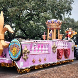 Op de afbeelding: Een roze en gouden paradewagen met een grote fleur-de-lis aan de voorkant. De wagen is versierd met gouden versieringen en heeft een rood gordijn aan de zijkant. De wagen wordt getrokken door een vrachtwagen met een grote band.