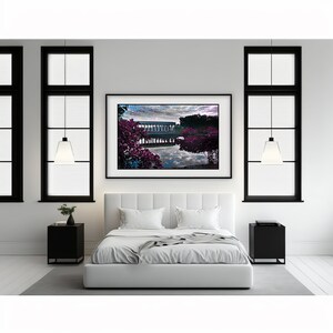 Op de afbeelding: Een wit bed met witte beddengoed en twee witte kussens in een slaapkamer met twee zwart geframede ramen en een zwart geframed beeld van een meer met een gebouw op de achtergrond en roze bloemen op de voorgrond.