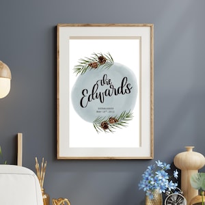 Può includere: Una stampa incorniciata con la scritta "the Edwards" in elegante corsivo, circondata da rami di pino e pigne su uno sfondo acquerello azzurro chiaro. La stampa è in una cornice di legno ed è esposta in una stanza dal design moderno.
