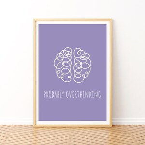 May include: A framed print with a light wood frame. The print has a lavender background with a white brain-shaped doodle and the words "PROBABLY OVERTHINKING" in white. The print is displayed on a wooden floor against a white wall.