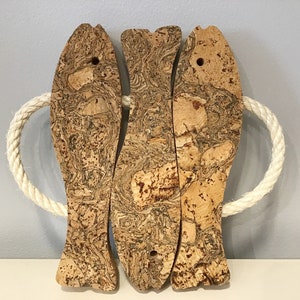 Può includere: Tre forme di pesce in sughero con un manico in corda bianca. I pesci sono in sughero naturale con un motivo a marmo marrone e beige.