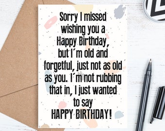 May include: A birthday card with the text "Sorry I missed wishing you a Happy Birthday, but I'm old and forgetful...HAPPY BIRTHDAY!" The card is on a white surface with a pen, a small container, and a brown envelope.